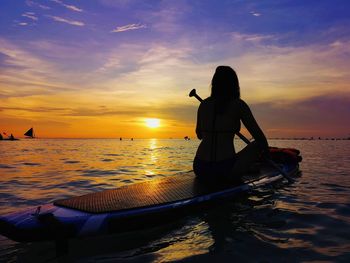 Silhouette woman sitting on paddleboard in sea against sky during sunset