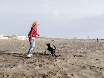 Full length of woman playing with dog on sand at beach against sky