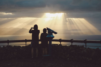 Silhouette couple standing by coin-operated binoculars with sea in background against cloudy sky