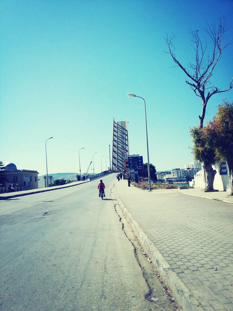 clear sky, blue, built structure, building exterior, architecture, copy space, tree, sunlight, street light, incidental people, transportation, the way forward, day, shadow, city, men, sky, road, outdoors