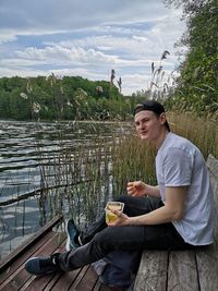 Side view of young man sitting by lake against sky