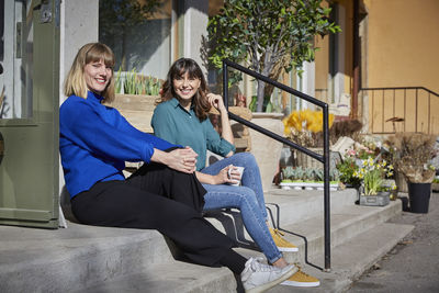 Full length portrait of smiling female partners sitting on steps outside store