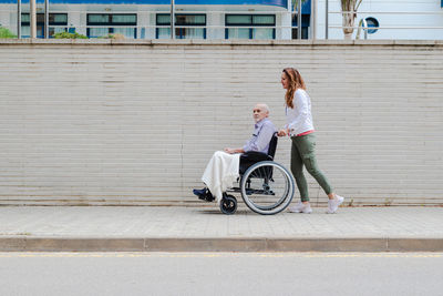 Side view of adult daughter pushing wheelchair with elderly father while walking together in city