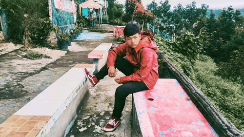 Side view of young man sitting on retaining wall against trees