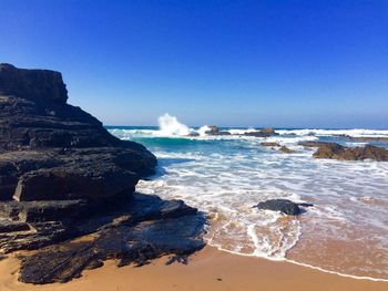 Scenic view of rocks by sea shore against clear blue sky