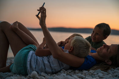 Rear view of people relaxing on shore against sky during sunset