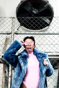 Portrait of woman wearing sunglasses while sticking out tongue against chainlink fence