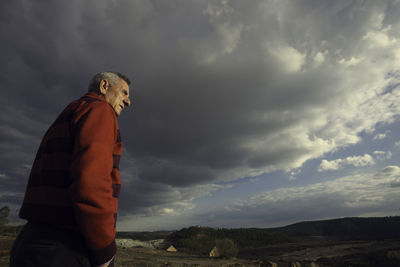 Side view of thoughtful senior man standing on field against cloudy sky