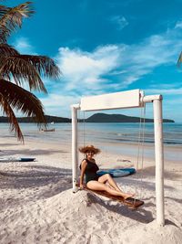 Full length of woman sitting on swing at beach against blue sky