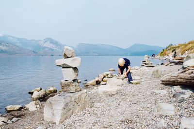 Boy picking up stone while standing on lakeshore against clear sky