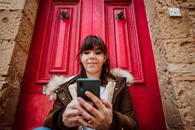 Portrait of woman holding mobile phone while standing against wall