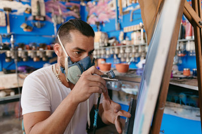 Side view of male artist in respirator using spray gun to paint picture on canvas during work in creative workshop