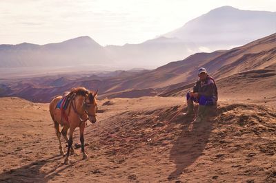 Full length of man sitting by horse at desert against mountains