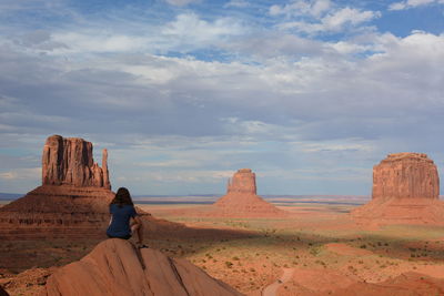Rear view of woman sitting on rock formations against cloudy sky