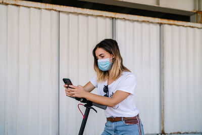 Side view of young female in casual outfit and sunglasses browsing mobile phone while standing with electric scooter near metal wall of urban building