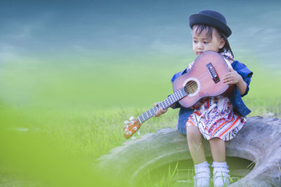 Cute girl holding ukulele while sitting on tire at grassy field