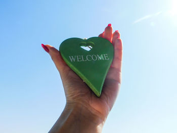 Cropped hand holding heart shape with welcome text against clear blue sky