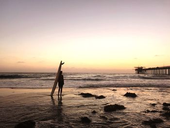 Man with surfboard standing at beach against sky during sunset