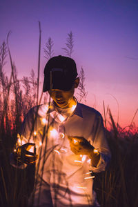 Young man holding illuminated string light while standing on field during dusk