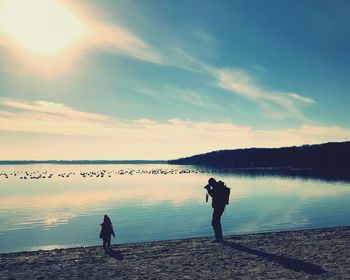 Silhouette father photographing daughter at lakeshore against sky during sunset