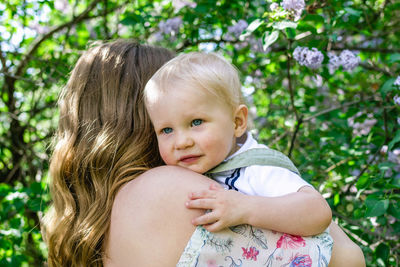 Portrait of mother with little son in her hands in park with lilac blossom tree, back view.