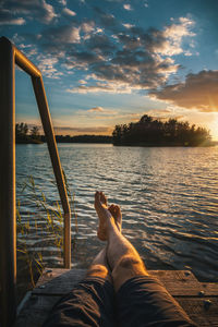 Low section of man sitting on lake against sky during sunset