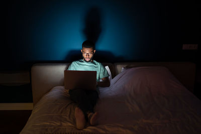 Young black man works on laptop lying in bed at night in dark looks attentively at screen.