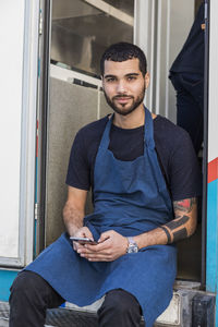 Portrait of confident young male entrepreneur sitting with smart phone at entrance of food truck