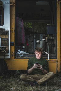 Full length of man sitting cross-legged while using smart phone against motor home during camping