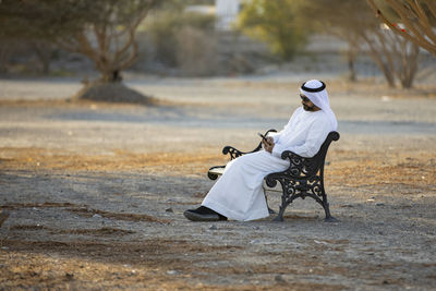 Full length of man sitting on bench using mobile phone outdoors