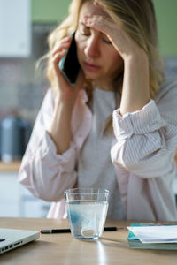 Depressed woman talking on phone while medicine diluting in glass