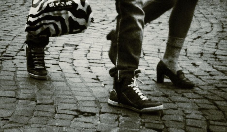 low section, shoe, person, high angle view, footwear, shadow, cobblestone, street, part of, sunlight, footpath, outdoors, human foot, day, standing, paving stone, close-up