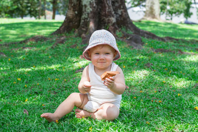 Adorable baby girl in white clothing and panama is playing with dry yellow leaves on grass. 