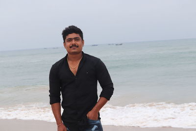 Portrait of young man standing at beach against sky