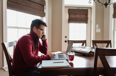 Man on cellphone working from home using a computer at a dining table.