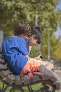 Side view of kids playing enjoying smart phone videos and games sitting on a park bench