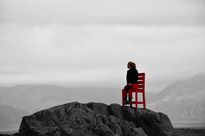 Young adult sitting on red chair on top of rocky hill during cloudy afternoon