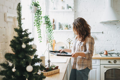 Attractive smiling woman with curly hair in plaid shirt bakes cookies at bright kitchen at home