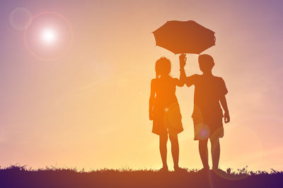 Silhouette siblings holding umbrella while standing on field against sky during sunset