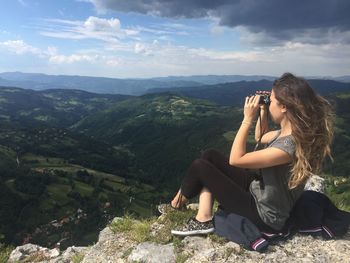 Woman looking through binoculars while sitting on cliff against sky