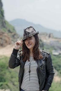 Portrait of smiling young woman wearing hat while standing on ground against sky