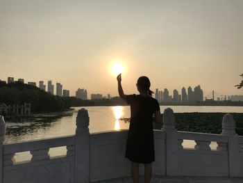 Rear view of woman gesturing towards sun while standing at balcony by river