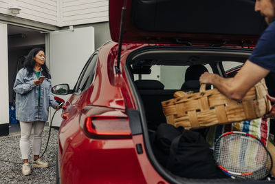 Woman holding smart phone while charging electric car near house