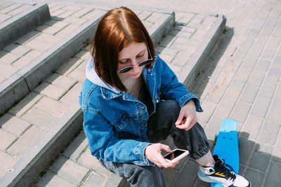 Young stylish woman walks on street with a skateboard and uses a smartphone