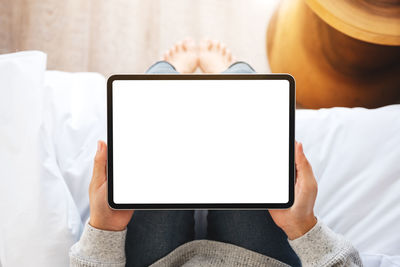 A woman holding black tablet pc with blank desktop white screen while sitting on a cozy white bed