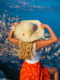 Young blonde hair girl in a hat sitting on the edge of a mountain with panoramic view of monaco