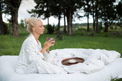 Beautiful woman 22-24 year old sitting in bed on white blanket drinking coffee outdoors over nature