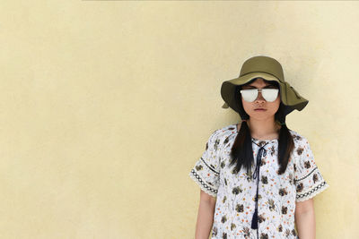 Portrait of young woman wearing sunglasses and hat while standing against wall