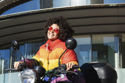 Woman wearing sunglasses and colorful jacket smiling while sitting on motorcycle