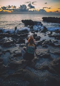 Rear view of woman sitting on beach against sky during sunset